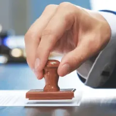 Mano con traje azul usa un sello de madera sobre un documento. El sello con firma es legal y oficializa el trámite.