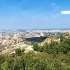 Panoramica delle **balze di Volterra**, con un casale in pietra in primo piano e dolci colline ondulate all'orizzonte sotto un cielo azzurro.