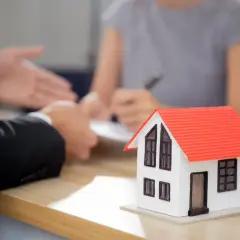 Model domu na stole, dłonie w garniturach, podpisująca dokumenty. Sytuacja sugeruje cofnięcie darowizny nieruchomości.
