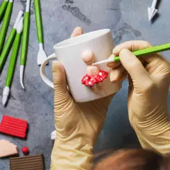 Mani guantate creano una bambolina con pasta fimo su una tazza. Tanti strumenti e colori per idee creative.