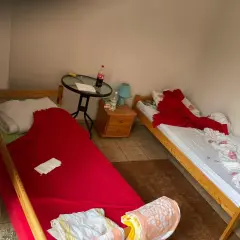 Pokój z dwoma łóżkami. Na jednym pościel po zmarłej osobie, przykryta czerwoną narzutą. Na drugim łóżku biała pościel z różowymi kwiatami i czerwony koc.