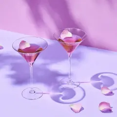 Dwa eleganckie drinki z martini rosato, ozdobione płatkami róż, unoszą się w delikatnym fioletowym świetle.