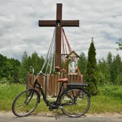 Rower przy kapliczce w Hajnówce, Białowieża. Drewniany krzyż z figurką Chrystusa i Matki Boskiej, otoczony zielenią i polnymi kwiatami.
