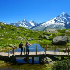 Zwei Wanderinnen auf einer Holzbrücke mit Blick auf den schneebedeckten Mont Blanc und eine grüne Berglandschaft.
