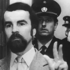 Odfried Hepp, mit Bart und im Anzug, hebt die Hand zum Friedenszeichen, umgeben von Polizisten.
