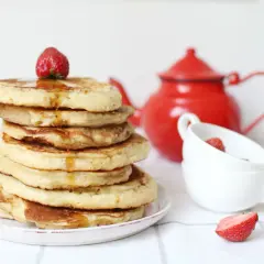 Pile de pancake sans gluten, nappés de sirop et garnis d'une fraise. Une théière rouge et des tasses complètent la scène.