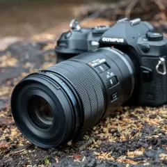 An Olympus camera with a Micro Four Thirds pancake lens rests on a bed of wood chips and dark soil, ready for an outdoor adventure.