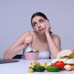 Une jeune femme s'ennuie en mangeant une salade, entourée de nourriture saine. Elle semble regretter de manger trop de fibres.