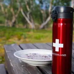Röd termos med schweizisk flagga och en skål gröt på ett träbord. Perfekt för en frukost vandring i naturen.