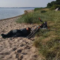 Osoba śpi na plaży obok roweru. W oddali widać wodę i drzewa. To dowód, że można spać na plaży.