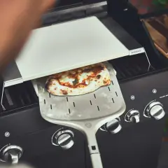 Frisch gebackene Pizza wird mit einem Pizzaschieber aus einem Gasgrill gehoben. Perfekt für den Sommer mit dem Pizza Gasgrill.