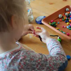 Dziecko z zaangażowaniem zajmuje się nawlekaniem koralików na sznurek, tworząc kolorową biżuterię.