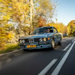 Błękitne BMW E9 z kierowcą jedzie jesienną drogą wśród kolorowych drzew.
