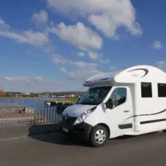 Un camping-car blanc roule sur un pont, avec une personne marchant à côté. Le ciel est bleu avec des nuages. Idéal pour la consommation camping-car.