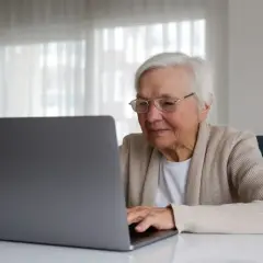 Uśmiechnięta starsza pani z okularami pracuje na laptopie, planując swoją emeryturę bez podatku.