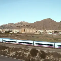 Tren AVE de Renfe, un moderno tren blanco con detalles morados, atraviesa un paisaje rural con montañas y edificios al fondo.