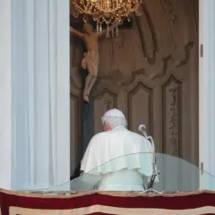 Der Papst steht auf dem Balkon, hinter ihm ein Kruzifix und ein Kronleuchter.