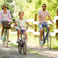 Rodzina cieszy się rodzinnym wyjazdem na **Mazury rowerem**. Radosna dziewczynka jedzie na fioletowym rowerze, a jej rodzice podążają za nią.