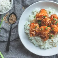 Ein Teller mit Reis und Hähnchen in süß-saurer Sauce, bestreut mit Sesam und Frühlingszwiebeln. Dieses Gericht hat Reis, Proteine und ist ein Genuss.