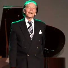 Uśmiechnięty Tadeusz Woźniakowski w czarnym garniturze, z niebiesko-białym krawatem, stoi na scenie obok fortepianu.