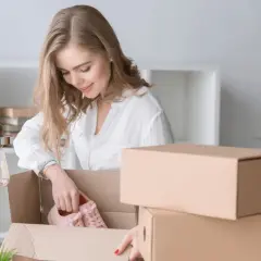 Kobieta rozpakowuje paczkę InPost, wyjmując z niej różowe buty. Obok stos kartonów.