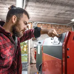 Mężczyzna w kratę reguluje panel sterowania nowoczesnego pieca na drewno. W tle worki z pelletem.