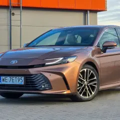 Brązowa Toyota Camry 2.5 Hybrid prezentuje swoje dane techniczne na tle industrialnego budynku.