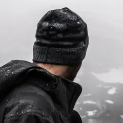Mężczyzna w czapce i ciemnej kurtce przeciwdeszczowej, na której widać krople wody, patrzy na mglisty krajobraz górski.