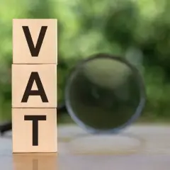 Drewniane klocki z literami VAT ułożone jeden na drugim, obok lupa. Analiza vat naliczony.