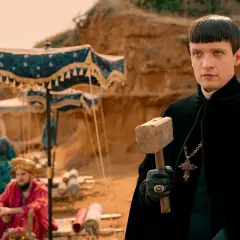 Młody mężczyzna w czarnym płaszczu, z krzyżem na szyi i młotem w dłoni, na tle obozowiska z "obsada 1670".