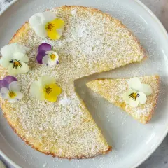 En saftig citron kladdkaka pudrad med florsocker och dekorerad med violer. En bit är uppskuren och en skål med grädde syns bredvid.