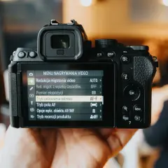 Dłoń trzyma aparat Nikon APS C, na ekranie widoczne menu nagrywania wideo z opcją "Tryb ustawiania ostrości" AF-F.