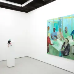 A miniature figure climbs a pedestal in a white-walled gallery, a vibrant painting of a diverse group at a meeting hangs nearby. NYC galleries for emerging artists showcase such intriguing pieces.