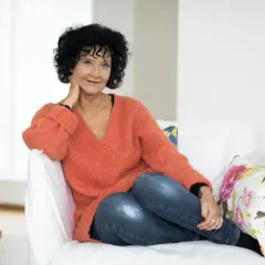 En kvinna med lockigt mörkt hår, klädd i en orange tröja och jeans, sitter avslappnat i en vit soffa. Hon ler mot kameran, med handen vid halsen. Lindell som skriver sina memoarer?