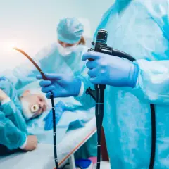 Lekarz w niebieskich rękawiczkach trzyma endoskop, przygotowując się do gastroskopii. Pacjent leży w tle.