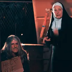 Una monja mira a un hombre con pelo largo y un cartel de "Ayúdame". Podría ser una escena de un sueño con monjas.