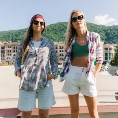 Dwie kobiety w letnich stylizacjach, jedna w koszuli w kratkę i jasnych rybaczkach, druga w zielonym topie i białych szortach, pozują na tle gór i budynków.