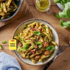 Makaron penne z pesto, boczkiem i bazylią na talerzu, obok słoiczek z pesto i tarką.