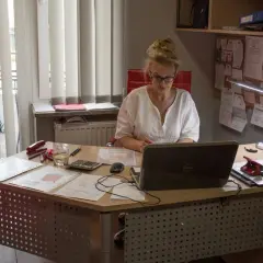 Kobieta w okularach pracuje przy biurku z laptopem, dokumentami i kalkulatorem.