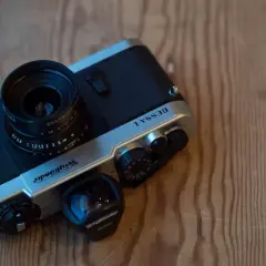 A silver and black Voiglander Bessa L camera with a lens attached and a separate 28mm lens nearby, resting on a wooden surface.