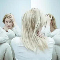 Kobieta z blond włosami w białej bluzce, trzymająca się za głowę, otoczona swoimi odbiciami w lustrze.