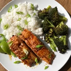 Två filéer av asiatisk lax med sesamfrön, serveras med ris, rostad broccoli och limeklyftor.