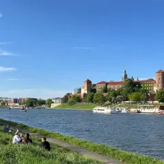 Słoneczny dzień nad Wisłą, ludzie odpoczywają na trawie, a w tle majaczy Wawel – symbol starego miasta Krakowa.
