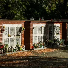 Kamienna ściana na urny z prochami, ozdobiona kwiatami i zniczami, otoczona jesiennymi liśćmi.