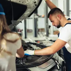 Mechanik z rękawiczkami bada silnik, szukając przyczyny usterki systemu zarządzania silnikiem. Obok stoi kobieta w czapce.