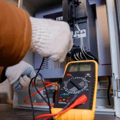 Elektryk w rękawiczkach sprawdza połączenia w szafie sterowniczej za pomocą żółtego multimetru. Pokazuje, jak używać multimetru do pomiarów.