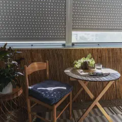 Przytulna loggia z bambusową ścianką. Drewniane krzesło z poduszką, okrągły stolik z napojem i roślinami. Idealne miejsce na relaks na balkonie.