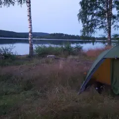 En grön campingtält står vid en sjö, med en skogklädd kulle i bakgrunden. En perfekt plats för äventyr längs 7 torpsleden.