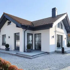 Nowoczesna, biała elewacja domu z brązowym dachem i dużymi oknami. Wokół zadbany ogród z brukowanym podjazdem.