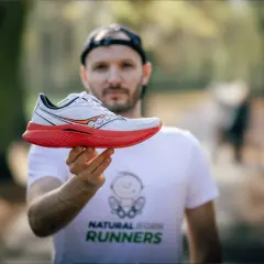 Mężczyzna w koszulce "Natural Born Runners" prezentuje biało-czerwony but biegowy Saucony. Saucony to znana firma z tradycjami.